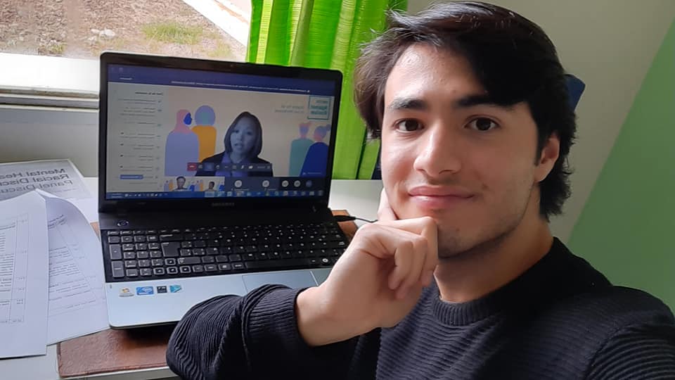 Es un selfi de Gabriel sonriendo, detrás está su computadora con la pantalla mostrando parte del evento.